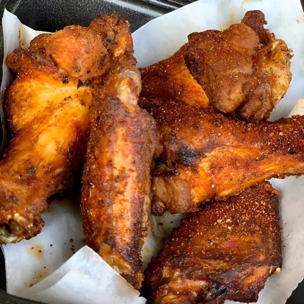 Close-up of several crispy, seasoned BBQ chicken wings and ribs in a black container with white paper.