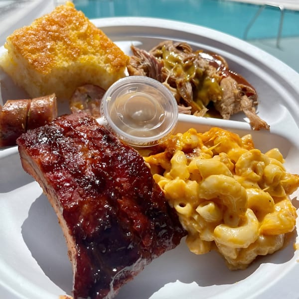 A white plate with Smoke BBQ ribs, pulled pork, mac and cheese, cornbread, and sausage.